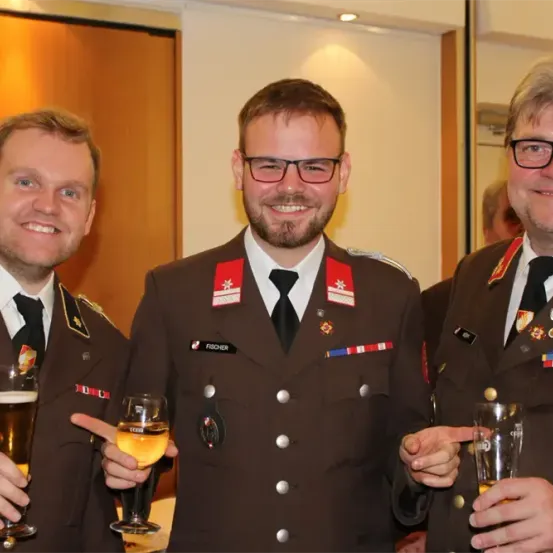 Drei Männer in Militäruniformen lächeln und halten Gläser mit Bier in der Hand. Sie stehen nebeneinander und scheinen für ein Foto zu posieren. Der Mann auf der linken Seite trägt eine Krawatte und eine Abzeichen.