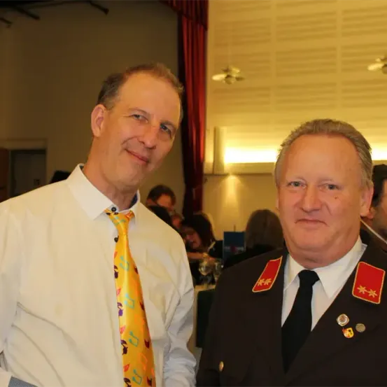 Zwei Männer stehen nah beieinander und lächeln für ein Foto. Einer trägt eine gelbe Krawatte, der andere ein schwarzes Uniform mit roten Schulterklappen. Hinter ihnen ist eine verschwommene Veranstaltung im Innenraum.