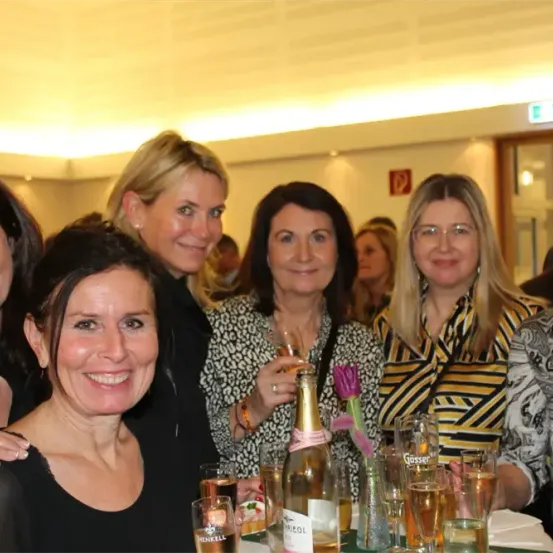 Eine Gruppe von Frauen lächelt und posiert für ein Foto bei einer gesellschaftlichen Veranstaltung. Sie sitzen um einen Tisch herum, auf dem Gläser, eine Flasche Wein und eine Blume stehen. Hinter ihnen stehen und unterhalten sich weitere Personen. Der Raum ist gut beleuchtet.