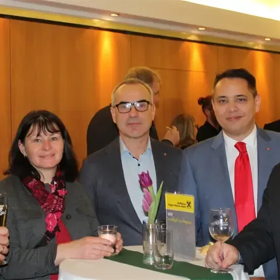 Fünf Personen lächeln in die Kamera. Drei Männer und zwei Frauen. Sie sind auf einer Party. Sie halten Getränke in den Händen. Vor ihnen steht ein Tisch.