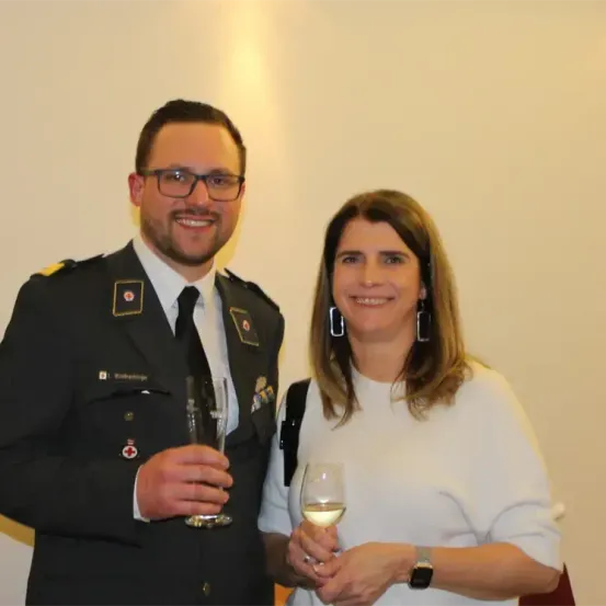 Ein Mann und eine Frau stehen nebeneinander und halten Weingläser in den Händen. Der Mann ist in Uniform mit einem Abzeichen und einem roten Kreuz. Die Frau trägt eine weiße Bluse.