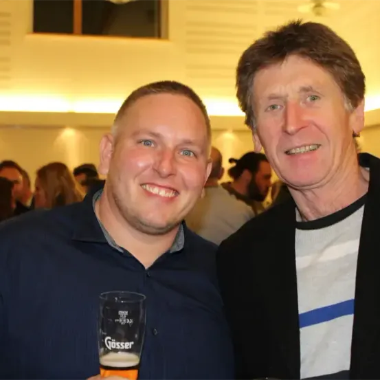 Zwei Männer lächeln und stehen bei einer sozialen Veranstaltung eng beieinander und halten ein Bier in einem Glas. Der Mann auf der linken Seite hat ein Glas Bier mit der Marke 'Gösser'.