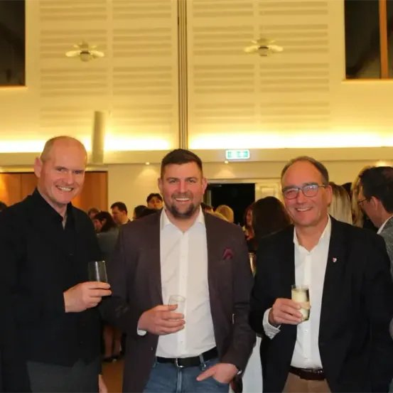 Vier Männer stehen zusammen in einem Raum und halten Getränke in den Händen. Sie tragen alle Anzüge und scheinen für ein Foto zu lächeln. Hinter ihnen sind weitere Menschen zu sehen, und der Raum hat ein modernes Innendesign.