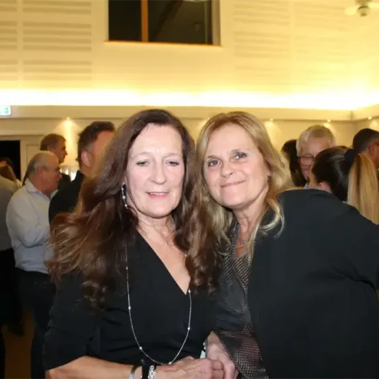 Zwei Frauen lächeln für ein Foto bei einer Veranstaltung und stehen zusammen mit anderen im Hintergrund.