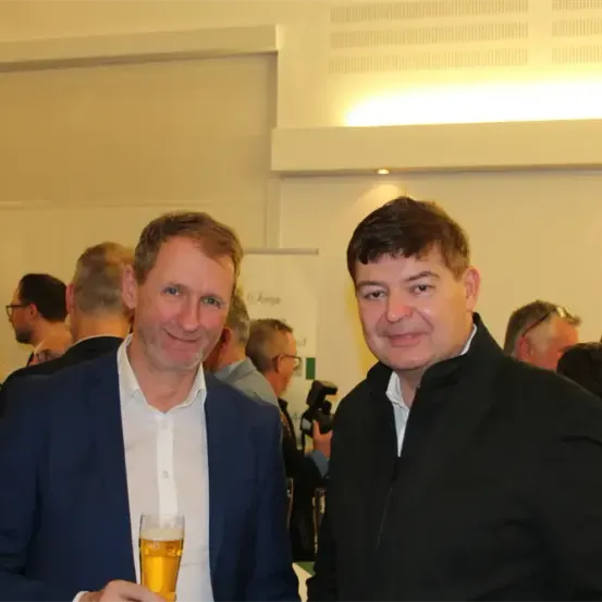 Zwei Männer lächeln und posieren für ein Foto bei einer Veranstaltung. Ein Mann hält ein Glas Bier. Im Hintergrund sind andere Teilnehmer zu sehen, einige mit Getränken.