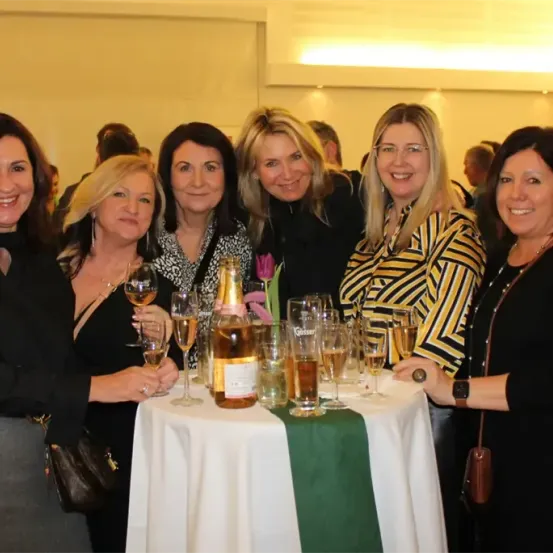 Eine Gruppe von Frauen steht um einen Tisch mit Weinglasern und einer Flasche und lächelt für ein Foto.