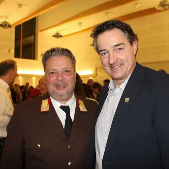 Zwei Männer stehen Seite an Seite und posieren für ein Foto bei einer Veranstaltung. Der Mann auf der linken Seite trägt eine Militäruniform.