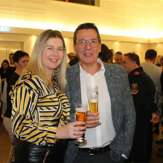 Ein Mann und eine Frau halten Gläser mit Bier in einer sozialen Umgebung, während andere im Hintergrund lächeln und umherschauen.