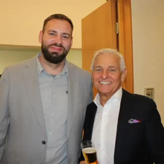 Zwei Männer lächeln und posieren für ein Foto, einer hält ein Glas Bier in der Hand und steht vor einer Holztür.