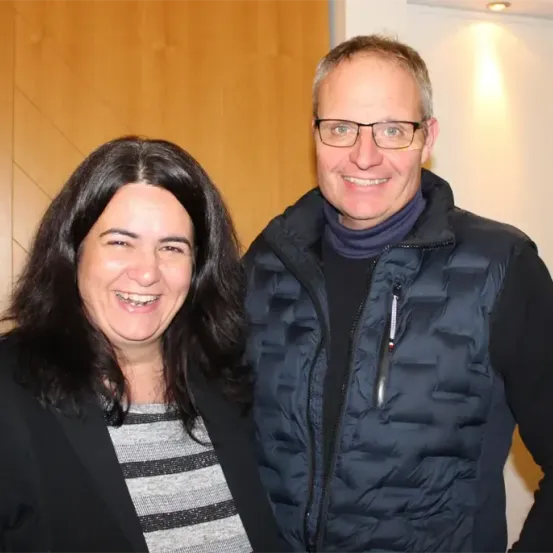 Ein Mann und eine Frau stehen lächelnd vor einer Wand. Die Frau hat lange dunkle Haare und der Mann trägt eine Brille und eine blaue Jacke.