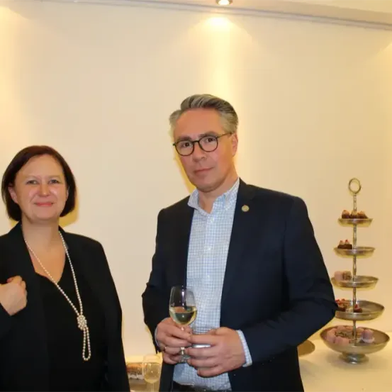 Ein Mann und eine Frau stehen nebeneinander und lächeln. Der Mann hält ein Glas Wein. Hinter ihnen steht ein Tisch mit einer Dessertpyramide und einem Weinglas.