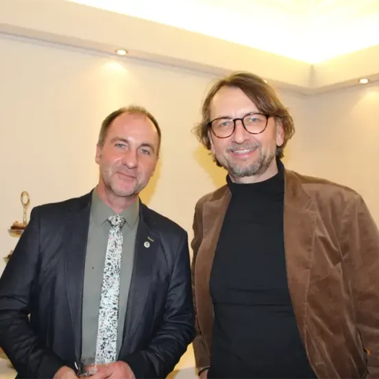 Zwei Männer stehen lächelnd in einem Raum mit weißer Wand und Decke. Der Mann auf der linken Seite trägt einen Anzug und eine Krawatte, der Mann auf der rechten Seite trägt eine Brille und eine braune Jacke. Hinter ihnen steht ein Pokal auf einem Tisch.