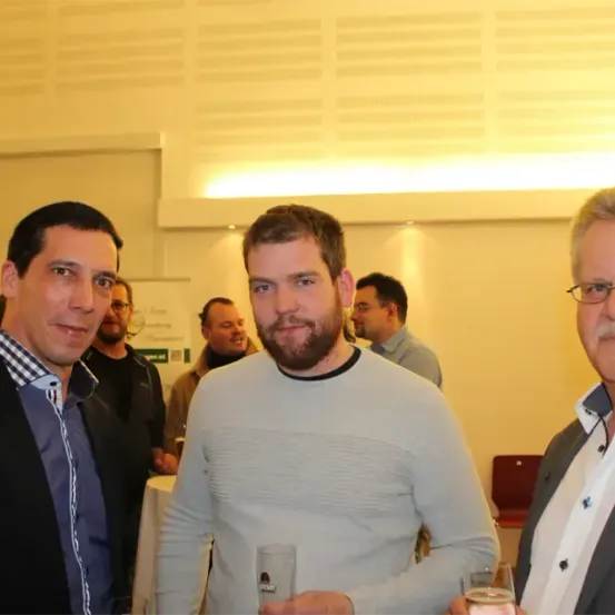 Drei Männer stehen in einem Raum und halten Getränke in den Händen. Der Mann in der Mitte hat einen Bart und trägt einen hellgrauen Pullover. Der Mann auf der linken Seite trägt ein kariertes Hemd. Der Mann auf der rechten Seite trägt eine Brille.