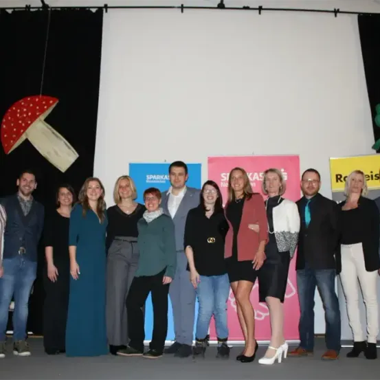 Eine Gruppe von Menschen steht vor einer weißen Wand mit drei Bannern dahinter. Sie scheinen für ein Foto zu posieren.