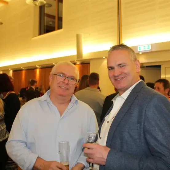 Zwei Männer, die lächeln und Getränke in der Hand halten, stehen nebeneinander in einer sozialen Umgebung. Dahinter scheint eine verschwommene Menschenmenge zu stehen oder zu sitzen und zu plaudern oder sich zu amüsieren.