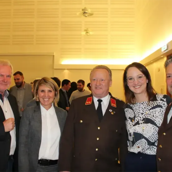 Eine Gruppe formell gekleideter Personen steht in einem gut beleuchteten Raum, lächelt und posiert für ein Foto. Zwei Männer in Anzügen stehen an den Seiten, eine Frau in einem gemusterten Shirt in der Mitte. Ein Mann in Uniform steht in der Mitte.