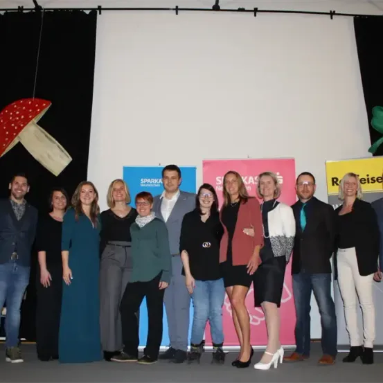 Eine Gruppe von Menschen, möglicherweise ein Team, steht zusammen auf der Bühne. Sie alle lächeln für die Kamera. Hinter ihnen sind Banner mit Text.