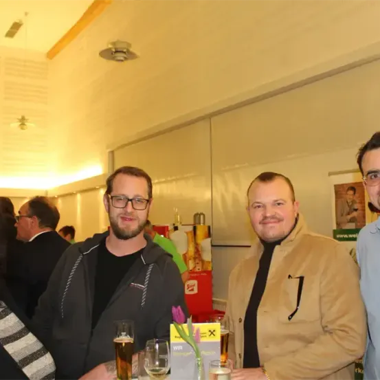 Drei Männer und eine Frau lächeln für ein Foto in einem Raum mit mehreren Personen im Hintergrund. Sie halten Getränke in der Hand und eine Blume ist im Vordergrund.