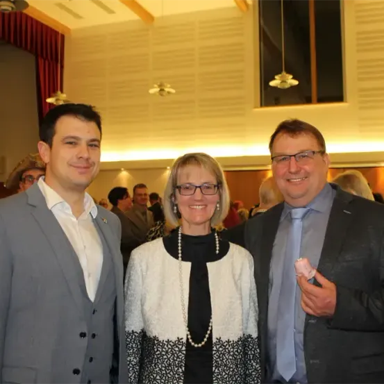 Drei Menschen lächeln und posieren für ein Foto bei einer Veranstaltung. Der Mann links trägt einen grauen Anzug, die Frau in der Mitte trägt einen weißen und schwarzen Pullover und der Mann rechts trägt einen blauen Anzug.