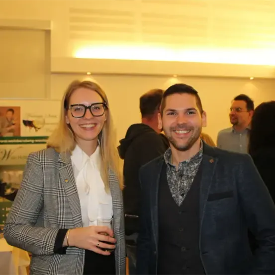 Eine Frau und ein Mann lächeln und stehen bei einer Veranstaltung nebeneinander. Die Frau hält einen Becher. Hinter ihnen stehen Menschen und unterhalten sich.