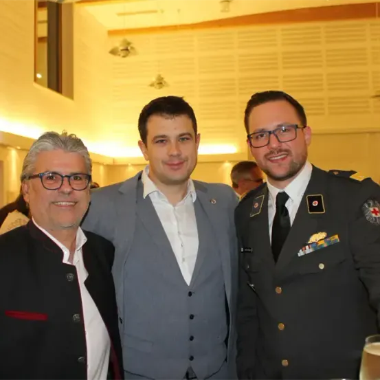Drei Männer stehen eng beieinander und lächeln für ein Foto. Der Mann links trägt eine Brille und einen dunklen Anzug. Der Mann in der Mitte in einem hellen Anzug steht unter einem hellen Licht. Der Mann rechts in Uniform hat ein Glas Wein.