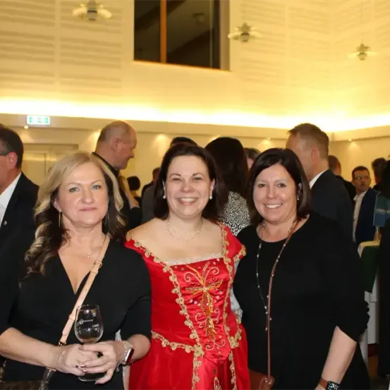 Drei Frauen lächeln in die Kamera; eine hält ein Weinglas. Dahinter stehen mehrere Menschen in formeller Kleidung in einem Raum mit weißen Wänden und dekorativen Deckenleuchten.