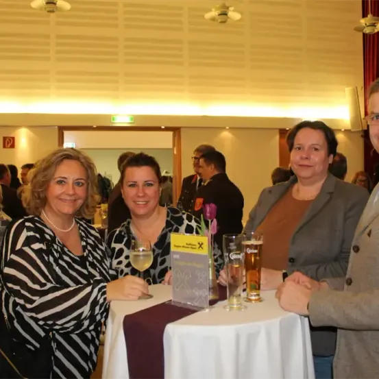 Vier Erwachsene sitzen lächelnd um einen Tisch. Eine Frau hält ein Weinglas. Der Tisch hat Biergläser und eine Speisekarte. Im Hintergrund stehen und sprechen mehrere Personen.