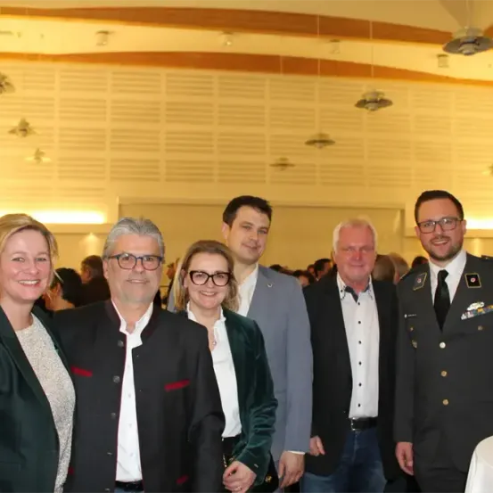 Eine Gruppe von Menschen, darunter zwei Frauen und drei Männer, posiert für ein Foto in einem formellen Rahmen. Ein Mann ist in Uniform, die anderen sind in formeller Kleidung. Der Hintergrund zeigt eine weiße Wand mit Lampen.