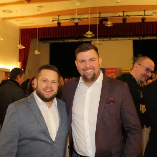 Zwei Männer stehen lächelnd für ein Foto bei einer formellen Veranstaltung, mit anderen im Hintergrund sichtbar.