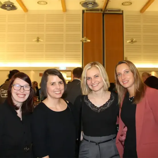 Vier Frauen in formeller Kleidung stehen eng beieinander und lächeln für ein Foto in einem Raum mit hängenden Lampen und Holzbalken.