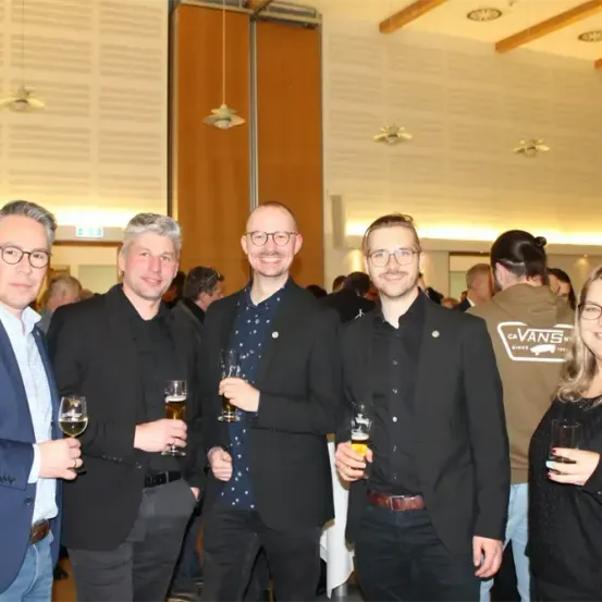 Fünf Erwachsene stehen eng beieinander, alle mit Brille und halten Getränke. Zwei Männer tragen Vans-Shirts. Eine Frau hält ein Glas.