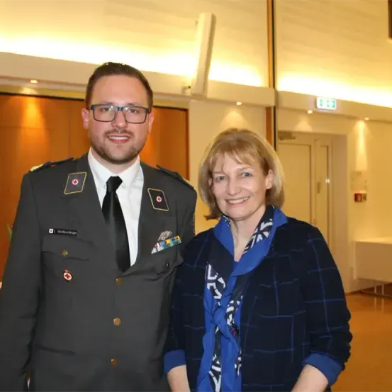 Ein Mann in Uniform und eine Frau in einem blauen Mantel stehen nah beieinander und lächeln, mit einer Holzwand und einem Tisch dahinter.