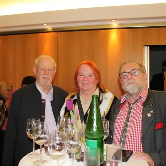 Drei Personen sitzen an einem Tisch mit Weinläsern und einer Flasche. Der Mann auf der linken Seite hat weiße Haare und trägt einen Anzug. Die Frau in der Mitte hat rote Haare und lächelt. Der Mann auf der rechten Seite trägt eine Brille. Hinter ihnen ist eine Holzwand.