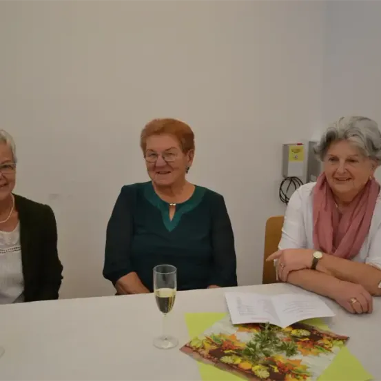 Drei ältere Frauen sitzen lächelnd am Tisch. Eine trägt eine Perlenkette, eine andere ein Tuch. Gläser und Weingläser sind vorhanden. Ein offenes Buch und ein blumengemusterter Stoff liegen auf dem Tisch.