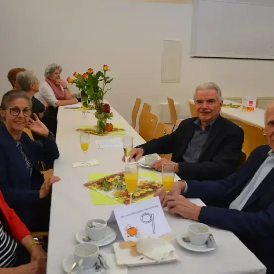Eine Gruppe von Menschen, wahrscheinlich bei einem Treffen, sitzt um einen langen Tisch herum. Sie haben Tassen, Teller und eine Blumenvase auf dem Tisch. Ein Mann lächelt und hält ein Glas. Der Tisch hat ein Schild mit der Nummer 9 und der Raum hat weiße Wände.