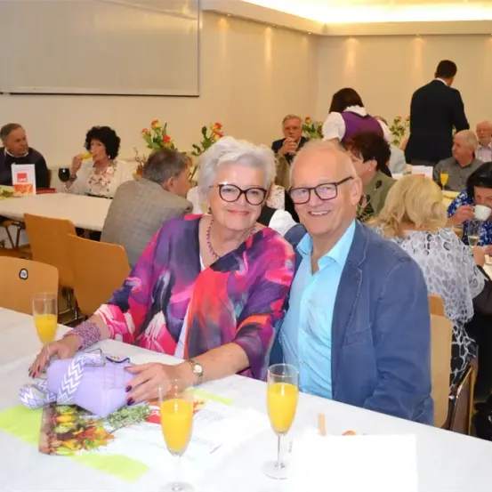 Eine Gruppe älterer Menschen, darunter ein älteres Paar, sitzt um Tische herum, wahrscheinlich bei einem gesellschaftlichen Treffen. Sie alle lächeln und scheinen ihre Getränke zu genießen.