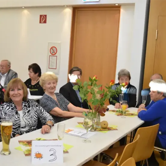Eine Gruppe älterer Menschen, möglicherweise bei einem Familientreffen, sitzt um einen Tisch herum. Eine Nummer-3-Karte, ein Blumenvase und Gläser stehen auf dem Tisch. Ein Schild mit einem Rauchverbotssymbol ist an der Wand angebracht.