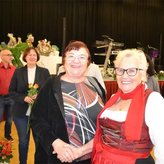 Zwei lächelnde Frauen mit Brillen schütteln sich die Hände. Dahinter steht eine Frau mit einem Blumenstrauß neben einem Mann in einem roten Hemd. Im Hintergrund befinden sich Pflanzen, Musikinstrumente und ein Tisch mit Blumen.