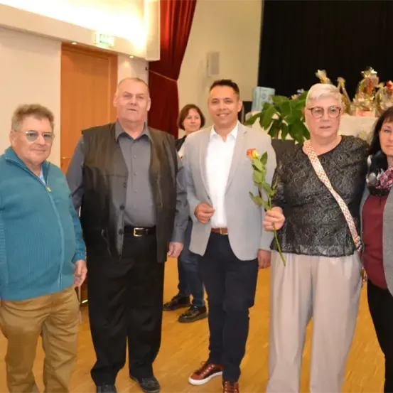 Fünf Menschen stehen in einem Raum zusammen. Eine Frau hält eine Rose. Eine andere Frau trägt eine Brille und einen Schal. Ein Mann trägt eine Weste.