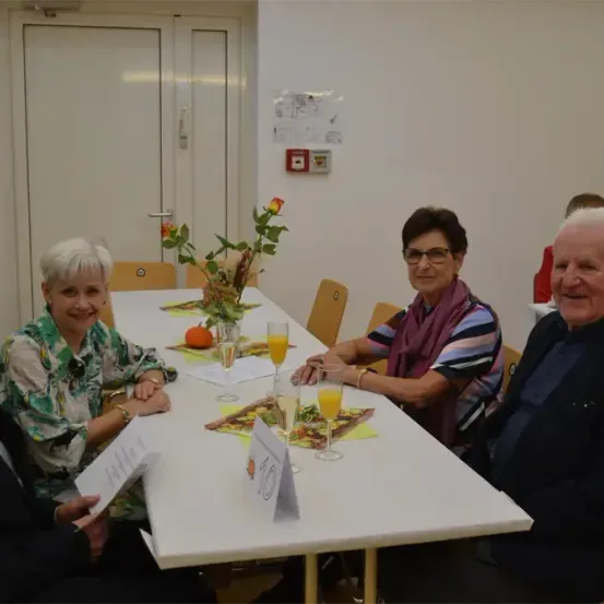 Vier ältere Menschen, zwei Männer und zwei Frauen, sitzen an einem langen Tisch mit Getränken und Essen und lächeln. Eine Blumenarrangement steht in der Mitte des Tisches.