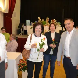 Vier Erwachsene stehen im Innenraum und lächeln für ein Foto. Die Frau in der Mitte hält ein Weinglas und einen Strauß Rosen. Ein Bühnenbereich mit verschiedenen Objekten befindet sich dahinter. Links steht ein Tisch mit Blumen.