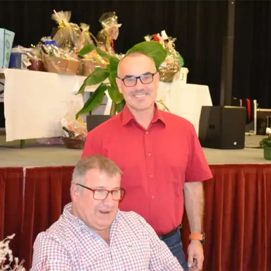 Zwei lächelnde Männer, einer steht auf der Bühne mit einem Tisch und Blumen hinter ihm, der andere sitzt auf einem Stuhl.