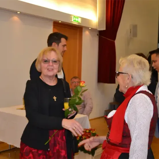 Eine Frau in einem roten Kleid hält eine Rose und unterhält sich mit einer anderen Frau in einem roten Mieder. Hinter ihnen stehen zwei Männer, einer in einem Anzug. Ein Tisch mit einer weißen Tischdecke und Blumen ist im Hintergrund.