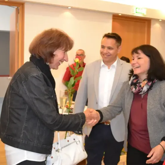 Drei Menschen lächeln und schütteln sich die Hände in einem Raum. Die Frau auf der linken Seite hält eine weiße Handtasche. Hinter ihnen steht ein Mann mit einer Rose und einem Glas Wein. Im Hintergrund steht ein weiterer Mann in der Nähe einer Tür.