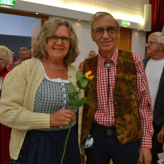 Ein Paar steht lächelnd nebeneinander. Die Frau hält eine Blume. Sie sind in einem Raum mit anderen Leuten, in der Nähe einer Tür.