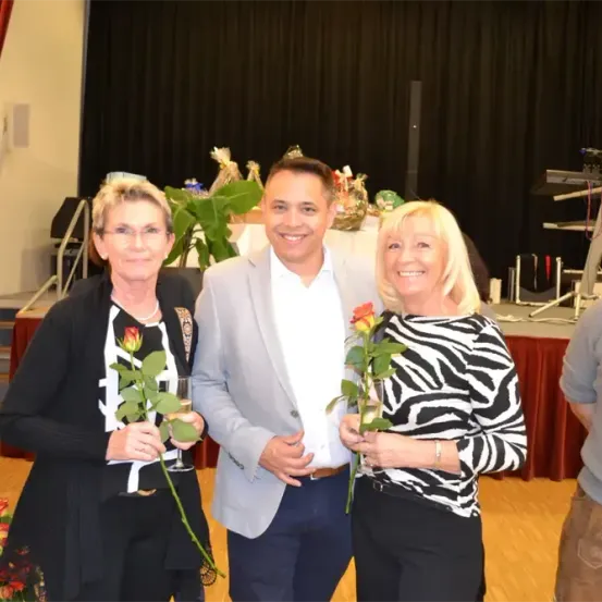 Drei Menschen stehen lächelnd für ein Foto zusammen. Zwei Frauen halten Rosen, eine in einem Zebra-Druck-Shirt, und ein Mann im Anzug. Ein Bühnenbereich mit einem Tisch und Pflanzen steht dahinter.