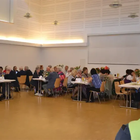Eine Gruppe von Menschen sitzt an Tischen in einem Saal, einige mit Getränken und andere mit Essen. Der Raum hat Holzböden, weiße Wände und eine Decke mit Lampen.