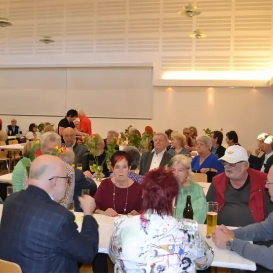 Ein Speisesaal voller älterer Menschen. Die meisten von ihnen sitzen um einen Tisch mit Blumen und Gläsern. Einige von ihnen tragen Hüte, Brillen und Halsketten.