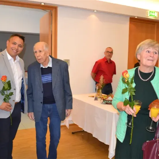 Drei Personen stehen in einem Raum; zwei Männer und eine Frau, halten Rosen und Gläser. Ein Tisch mit Essen ist hinter ihnen.