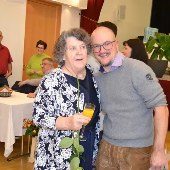 Eine lächelnde ältere Frau in einem gemusterten Kleid und ein Mann mit Brille und grauem Pullover stehen eng beieinander und halten ein Glas Orangensaft. Hinter ihnen steht ein Tisch mit Blumen und einer sitzenden Frau. Im Hintergrund sind Menschen, eine Tür und eine Pflanze zu sehen.